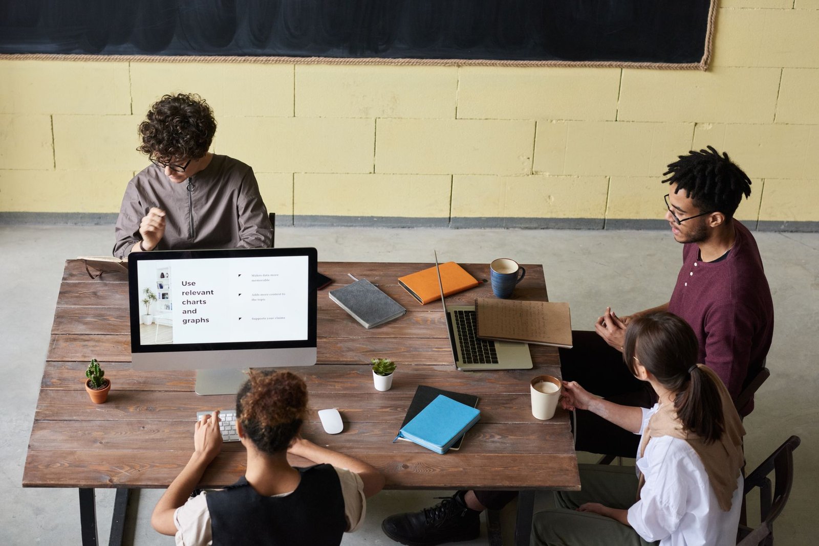 Four diverse colleagues engaged in a team meeting, working on laptops with coffee and notes, fostering collaboration. — How T